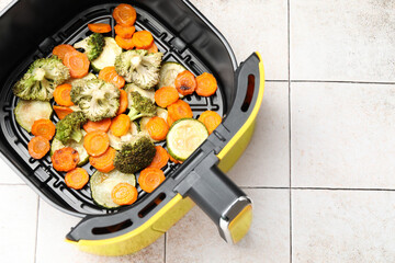 Cooked carrots, broccoli and zucchini in air fryer basket on white tiled table, top view. Space for text