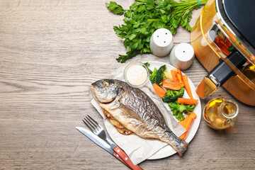 Tasty grilled fish, vegetables, sauce and air fryer on wooden table, flat lay. Space for text