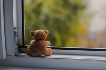 Sad brown teddy bear alone sitting on open window sill and looking out of home.