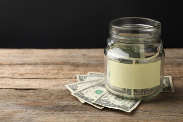 Jar with blank note and money on wooden table against black background. Space for text