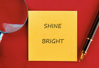 A vibrant yellow sticky note displaying the words shine bright is placed on a deep red surface. Next to it are a sleek pen and a magnifying glass, inviting creativity and inspiration