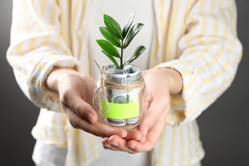 Woman holding jar with blank note, money and sprout on grey background, closeup