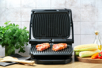 Electric grill with uncooked steaks, vegetables, utensils and herbs on wooden table