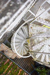 A high-angle view of a weathered, outdoor metal spiral staircase. The diamond plate steps provide an industrial texture. A concept of utilitarian architecture and urban exploration.