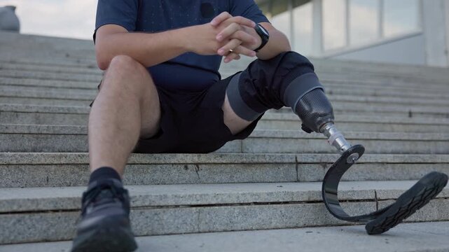 Male amputee athlete with running blade prosthetic resting on outdoor stairs after jogging, close up shot. Disability, sport, and recreation concept.