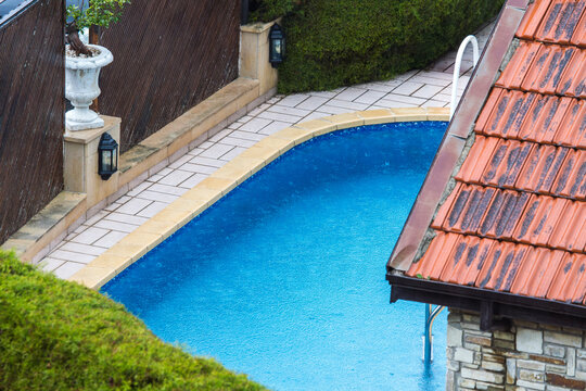 Rain creates ripples on the vibrant blue water of a backyard swimming pool, seen from a high angle. The scene captures a quiet, moody moment contrasting the wet terracotta roof with the tranquil water