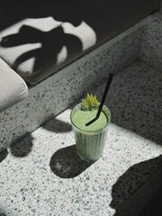 Green smoothie with apple garnish in glass with straw, sunlight and shadow on terrazzo