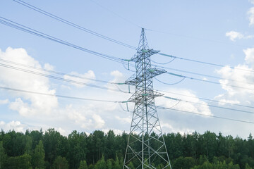 Tall electricity pylon with power lines extending across a blue sky with white clouds above a green forest. Energy transmission and infrastructure concept.