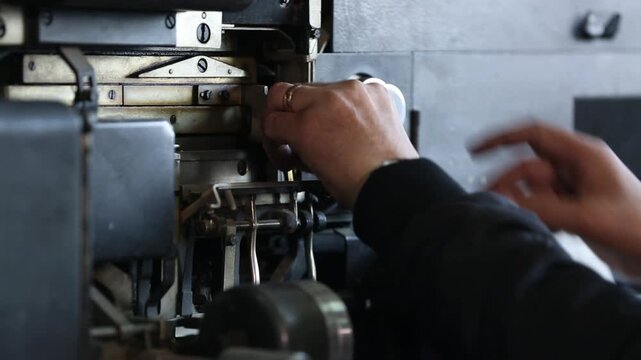 Skilled worker adjusts and maintains complex vintage printing press or typesetting machine. Detailed close-up of hands on intricate industrial components and levers.