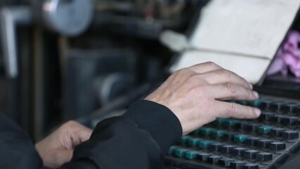 Skilled hands operate a vintage industrial keyboard, likely a Linotype machine, inputting data from handwritten notes. This represents historical printing technology and dedicated factory work.