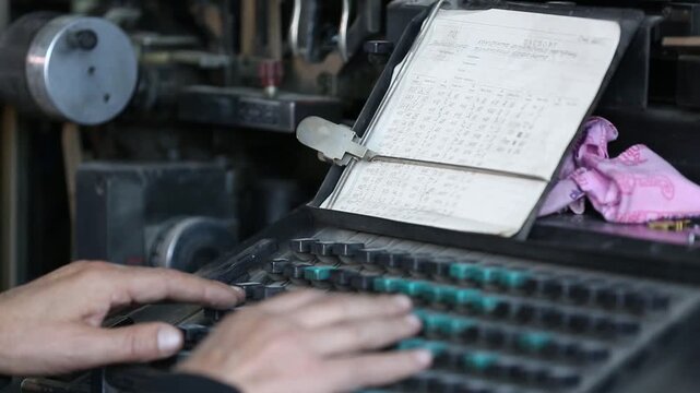 Skilled hands operate a vintage industrial keyboard, likely a Linotype machine, inputting data from handwritten notes. This represents historical printing technology and dedicated factory work.