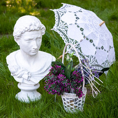 Elegant white classical bust, lace parasol, and wicker basket with purple flowers on lush green grass &mdash; romantic, vintage-inspired garden scene perfect for weddings, decor, or lifestyle themes.