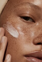 Fototapeta premium Close-up of person with freckles applying white cream to cheek. Macro shot skincare photography.