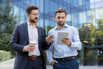 Two businessmen colleagues discussing digital data on a tablet device, standing outdoors with a...