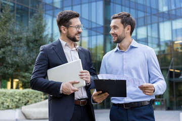 Two smiling adult businessmen, one holding a laptop and the other a clipboard with documents,...