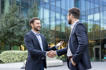 Two smiling businessmen shake hands outdoors in front of a modern corporate building, sealing a...