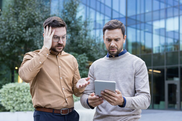 Two stressed businessmen looking at a digital tablet with worried expressions, reacting to negative...