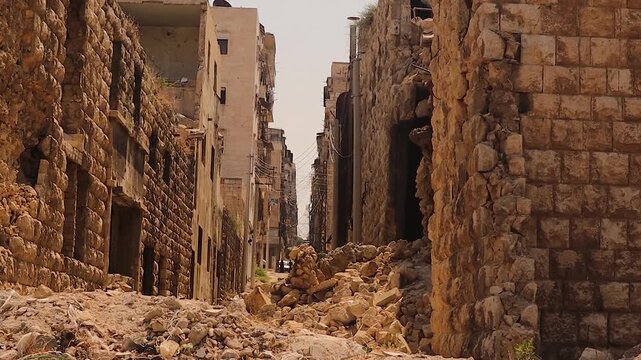 War damaged street in Aleppo, Syria, with destroyed stone buildings and rubble blocking the way. In the distance, people return, symbolizing resilience and the importance of reconstruction.