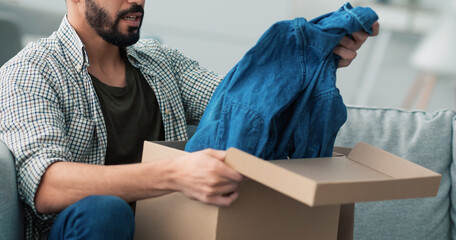 A man sits on a couch, excitedly unpacking a cardboard box. He pulls out a stylish denim shirt,...