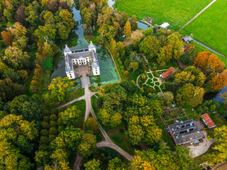 Aerial view of historic castle symmetrical twin towers and central hall framed by forest, moats, and tree lined path, with open fields and residential zone beyond under warm sunrise sky.