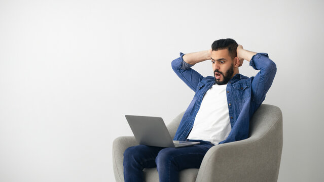 A young man sits in a modern chair, looking shocked and bewildered as he stares at his laptop. He is wearing a denim jacket over a white shirt, suggesting he is at home during a casual moment. - Powered by Adobe