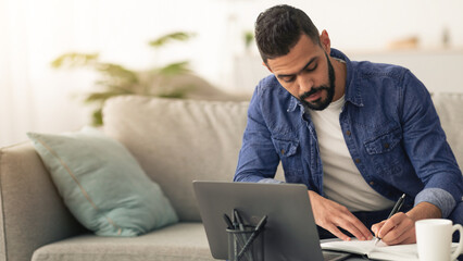 A young man focuses on his work at home, writing notes in a notebook. He sits comfortably on a...