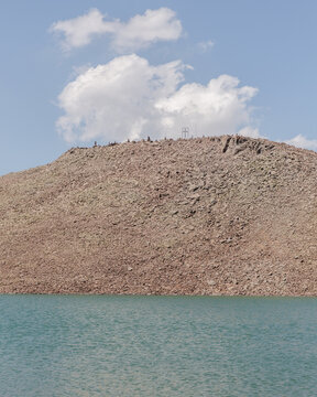 View of a rugged, sun-baked hill rising sharply from the still, turquoise waters under a bright sky dotted with fluffy white clouds, Lake Kari, Shirak Province, Armenia.