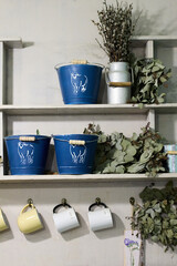 A delightful rustic kitchen shelf decorated with charming blue buckets featuring sheep, delicate dried eucalyptus, soft pussy willow branches, and welcoming hanging mugs, creating a cozy scene.