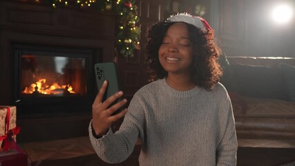 Merry Christmas. African woman in Santa hat having video chat in smartphone. Blogger in festive mood recording video in social network. Girl having video call with friends family celebrating Christmas - Powered by Adobe