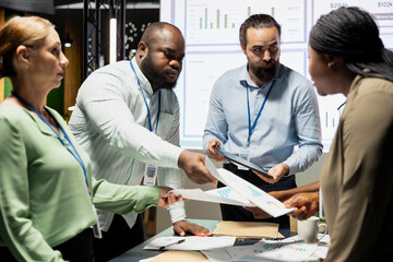 Multiethnic work team exchanging documents and using data visualization to forecast business goals in a quiet corporate strategy room. Staff members collaborating late at night.
