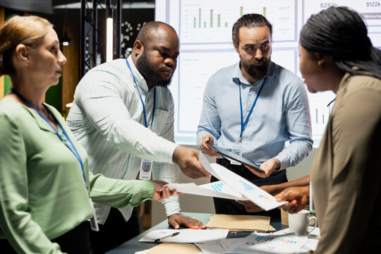 Multiethnic work team exchanging documents and using data visualization to forecast business goals in a quiet corporate strategy room. Staff members collaborating late at night.