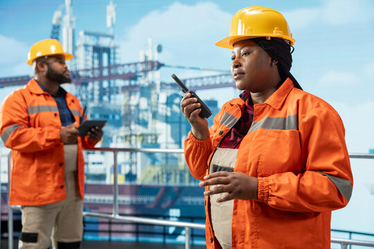 Offshore platform professional using hand portable radio device to coordinate workflow during operations. African american drilling rig worker speaking in walkie talkie, doing emergency procedures - Powered by Adobe