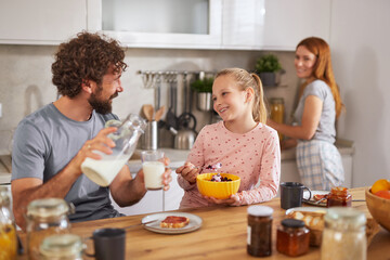 A father and daughter share a joyful breakfast in a modern kitchen. They engage in a lighthearted conversation while enjoying pancakes and milk, with a mother in the background.