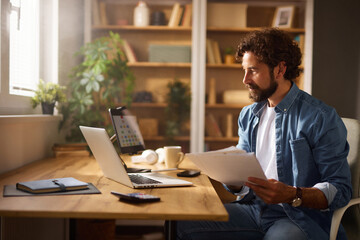 A man is engaged in work at a modern home office, seated at a wooden desk with a laptop and papers in front of him while surrounded by houseplants and shelves.