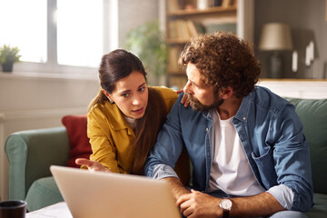 A couple appears to be having a serious conversation about financial problems while seated on a green couch in their cozy living room, using a laptop to review information.