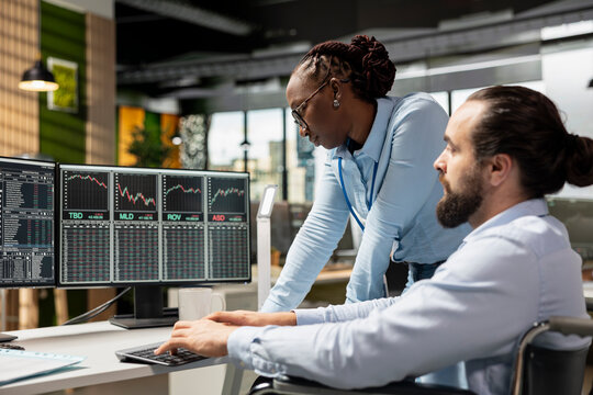 Risk analyst and coworker with disability in inclusive office using trading platform to review stock market volatility. Person with disability in wheelchair and colleague analyzing portfolio reports