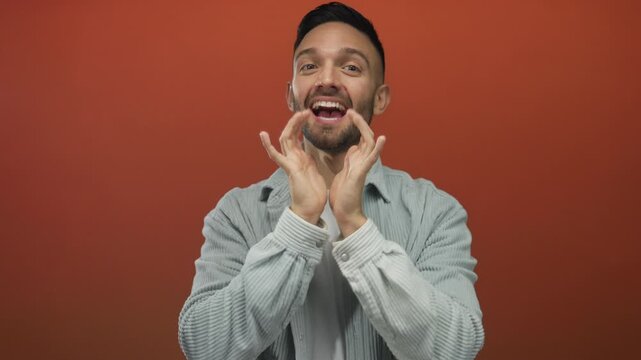 Young man enthusiastically shouting with hands framing face against vibrant orange background conveys excitement and energy.