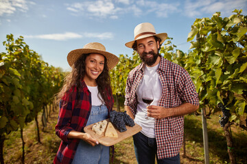 Two happy vineyard owners proudly present a wooden platter with grapes and cheese. Surrounded by rows of green vines, they celebrate their family business in wine production on a sunny day.