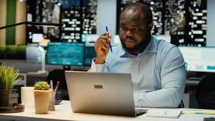 Black general manager envisioning a tactical solution and playing with pen, working late alone in office. Guy engaging in critical thinking, evaluating options and solutions for dilemma.