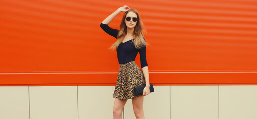 Beautiful stylish young woman in leopard print skirt with clutch bag standing on city street