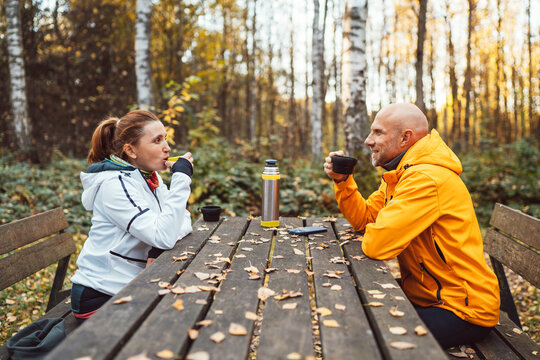 Middle-aged couple resting after walk enjoying hot tea from thermos at wooden picnic table in autumn forest. Active lifestyle, outdoor relaxation cozy moment, connection with nature during fall season