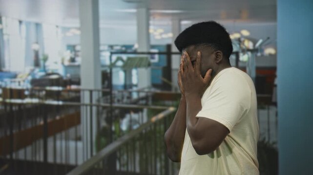 Young man covers face with both hands beside hotel mezzanine railing in building interior; anxiety isolation.