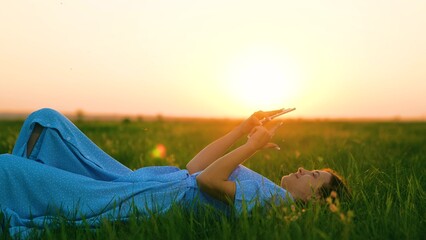Girl lies on green grass, uses smartphone. Happy teenager relaxes with gadget on green lawn at sun park. Technology in hands of younger generation. Favorite gadget for outdoor recreation. Gadget