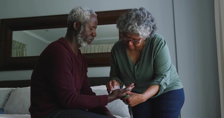 Performing senior woman checking man's blood glucose on sofa in living room, with meter and lancet