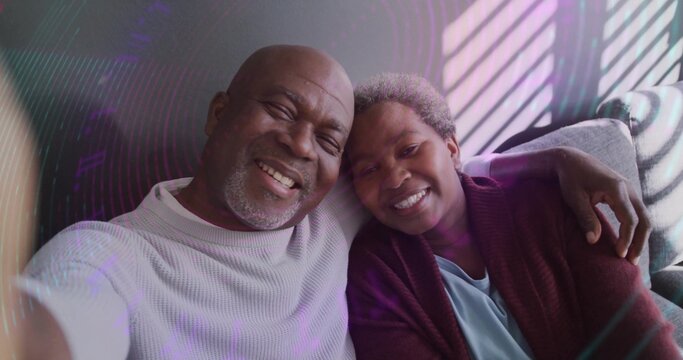 Smiling senior couple leaning together taking selfie on light sofa, with window blinds shadows