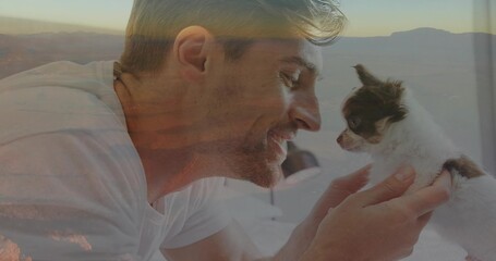Leaning man wearing t-shirt holding small brown white puppy in bedroom, with white bedding and lamp