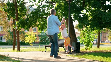Happy dad son are spinning in city park. Happy family, father little son, are playing outside park summer. Dad spinning child around, holding his hands little boy is laughing. People park, parent kid