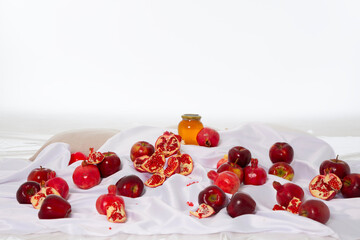 Horizontal photo. Red ripe fresh juicy fruits, apples, pomegranates and glass jar of honey on white textile fabric tablecloth. Decor for Rosh Hashanah - Jewish New Year holiday. Concept of health food