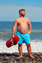 Vertical travel photo. Young adult tanned fat white man with stubble, beard, blue shorts, big belly stands on pebble sea coast looks side. Lifeguard holds lifesaving buoy. Sun day. Tourism, trip, calm