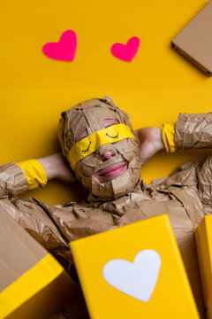 Surreal woman resting on yellow background wrapped in tape with drawn eyes &ndash; conceptual art about love, perception, and playful illusion